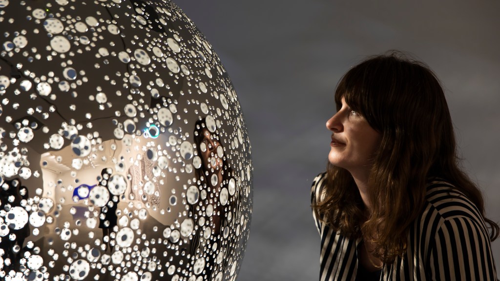 a woman looking at a shiny round reflective sculpture in a gallery