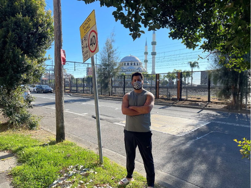 A man with crossed arms and face mask stands in front of a road with a mosque in the background