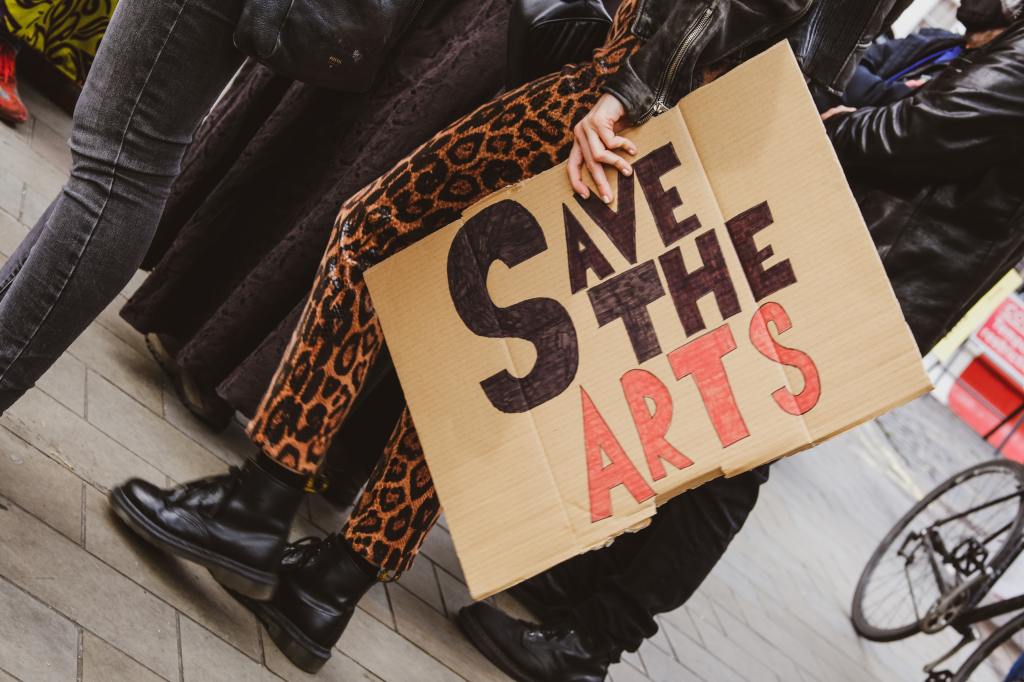 Person in leopard-print pants holds a sign saying Save the arts