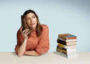 A woman hold a book over her face with a stack of books to one side