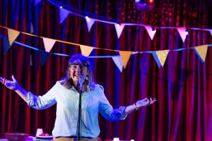 A woman holds up her hands in celebration with bunting over her and red curtain behind her