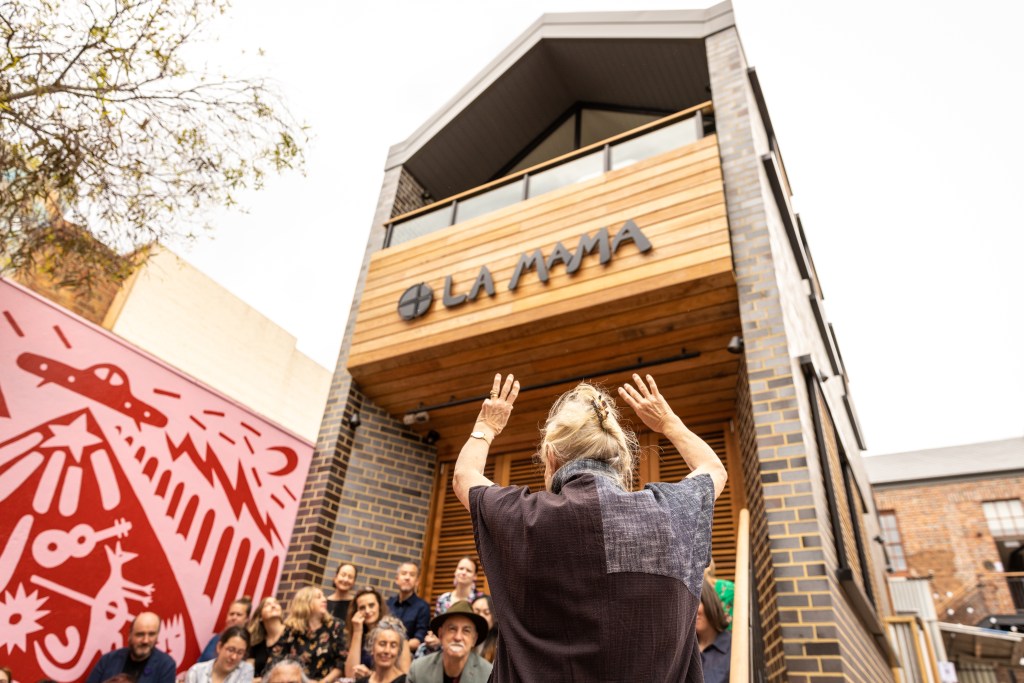 A woman in front of a small crowd raises her hands towards a building with a sign reading La Mama