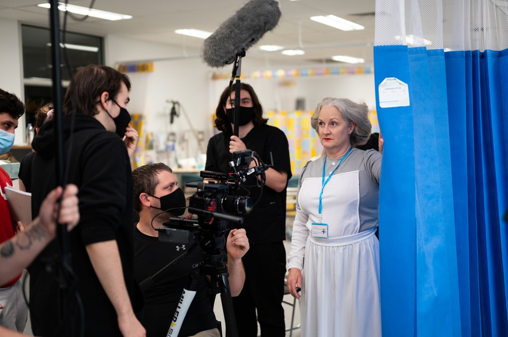 A film set in a hospital with a nurse and several camera operators surrounding her.