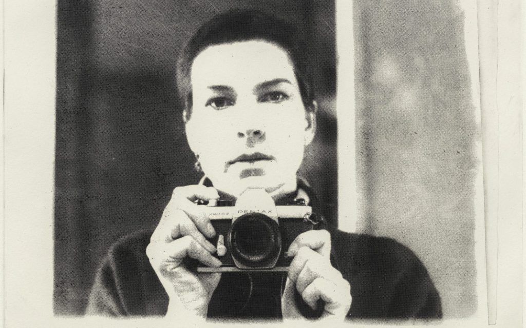 a black and white photo of a woman with a close shaven head looking at the camera, holding a camera