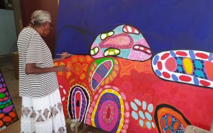 an elderly female Aboriginal artist painting a large canvas work