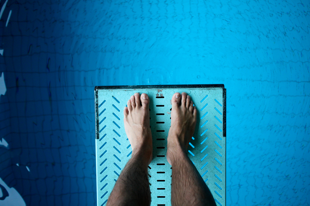 A pair of feet at the edge of a diving board with blue water below.
