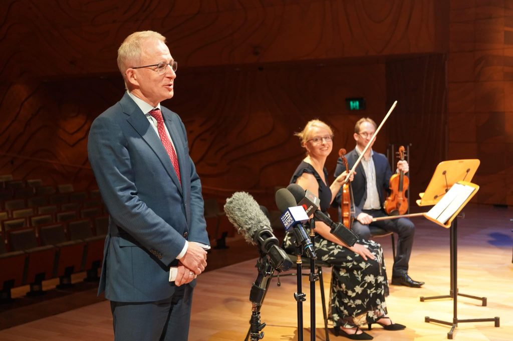 Paul Fletcher at a microphone with two violinists in the background