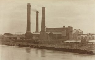 A sepia-toned photograph of the old Richmond Power Station by the banks of the Yarra, taken in the 1920s. Thick smoke blows from three tall chimneys.
