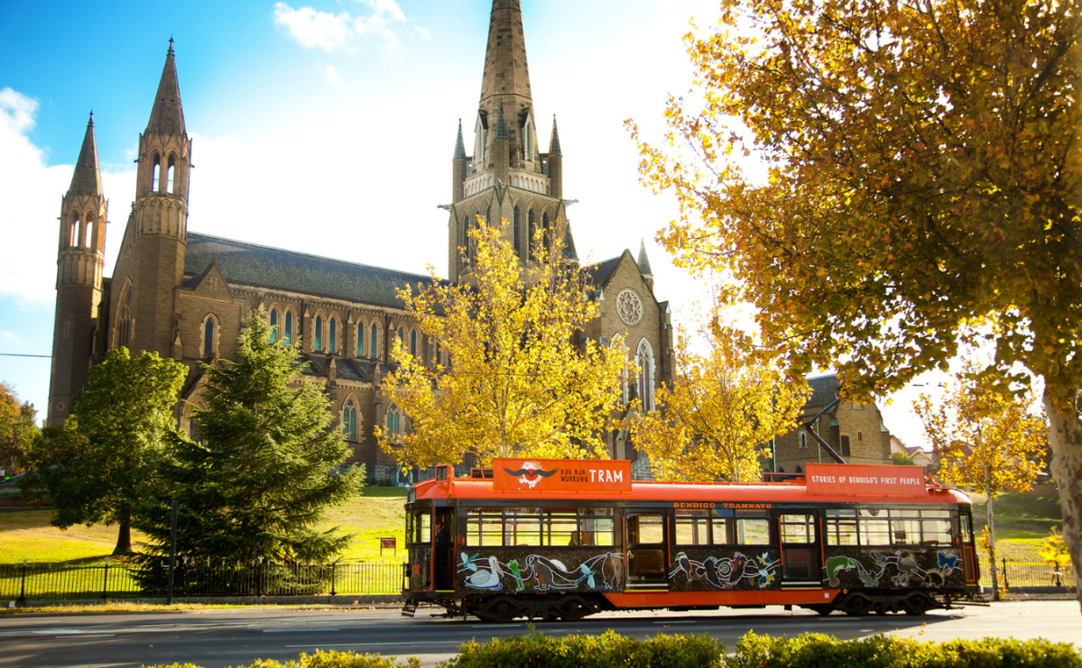 Autumn leaves frame a view of a cathedral against a cloud-scattered sky as a tram passes in the foreground.