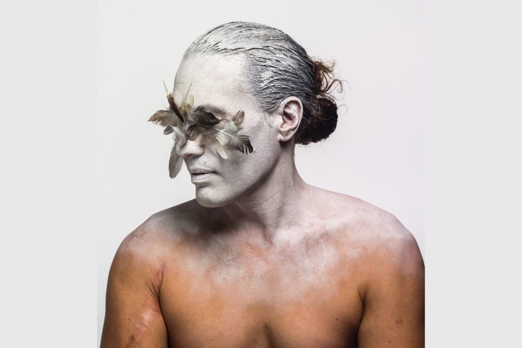 a male performer with bare chaest covered in white powder with leaves over his eyes