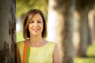 A woman wearing a tellow dress with orange and green stripes (Jo Dyer) stands beside a gum tree.