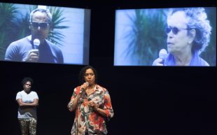 two indigenous actors (Kylie Doomadgee and Rachael Maza) on stage, one is speaking into a microphone, backgrounded by screens showing Indigenous people holding microphones