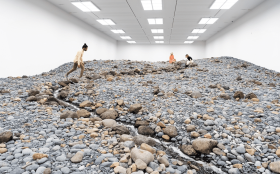 A large white gallery room with fluorescent lighting is filled with stones and rocks, plus a water course - an installation called Riverbed.