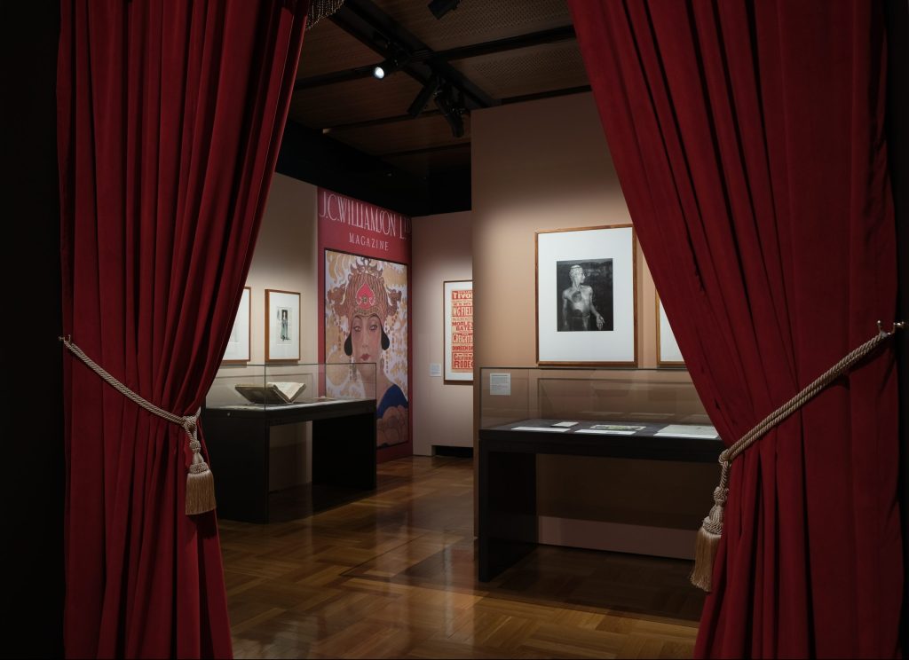 Red velvet curtains are drawn aside to provide a glimpse of an exhibition, with posters and other artefacts visible on walls and in display cases.