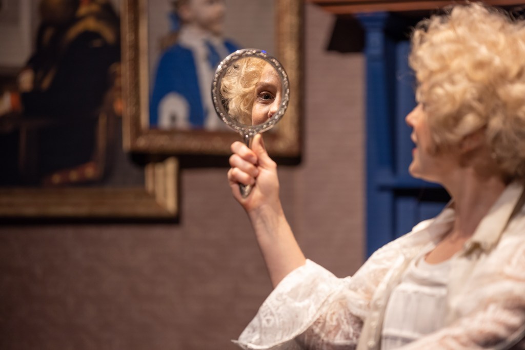 Erin Jean Norvill in a blonde wig as Dorian Gray gazes at himself/herself in a hand-held mirror.