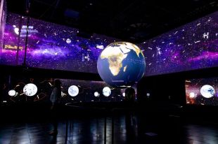 A dark exhibition space with a series of lit up globes hanging in it.