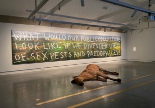 A typical 20th century landscape painting hangs on a white wall, with capitalised letters written across, “what would our public collections look like if we divested them of sex pests and paedophiles?” A sculpture of a realistic dead horse lies at its feet.