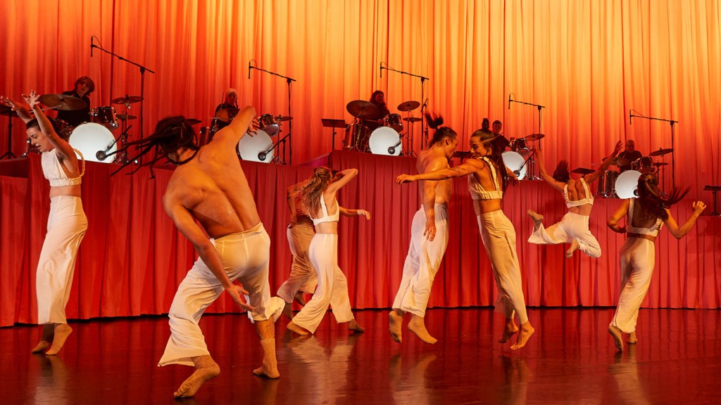 A group of male and female dancers dressed in white dance against an orange cloth blackground.