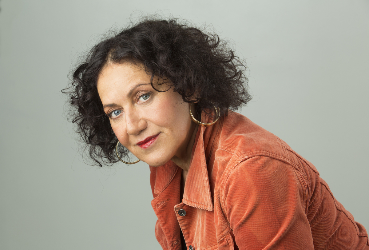 Comedian Rachel Berger looks into the camera. She has wavy brown hair and wears large hoop earings and an orange jacket.