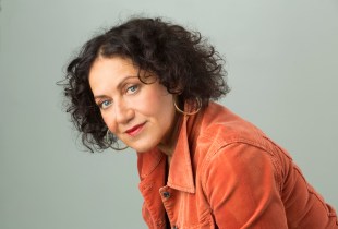 Comedian Rachel Berger looks into the camera. She has wavy brown hair and wears large hoop earings and an orange jacket.