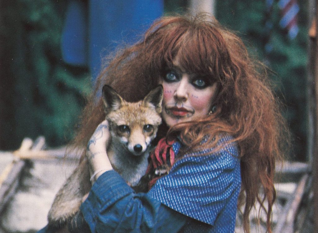 A red-haired woman, her eyes dark with kohl, holds a fox and stares into the camera.