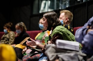 A woman with dark bobbed hair and wearing a green knitted vest leans forward, her hands animated, photographed mid-discussion. Other people sit behind and around her though they are not in focus.