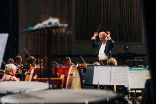 A grey-haired man in formal wear and glasses stands at the podium with both hands raised, a conductor's baton in his right hand. Members of the orchestra sit around him.