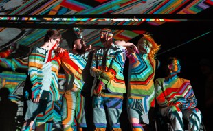 Brightly coloured projections wash over a group of five people standing and sitting close together, making their features hard to distinguish.