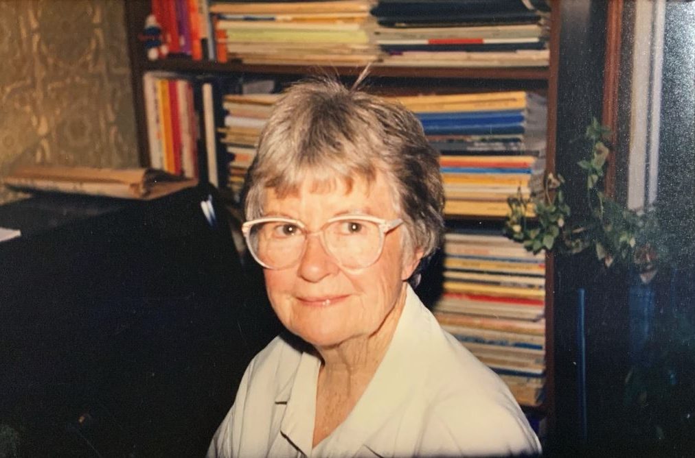 A grey-haired woman wearing a white shirt and white-framed glasses sits in front of a crowded bookcase.