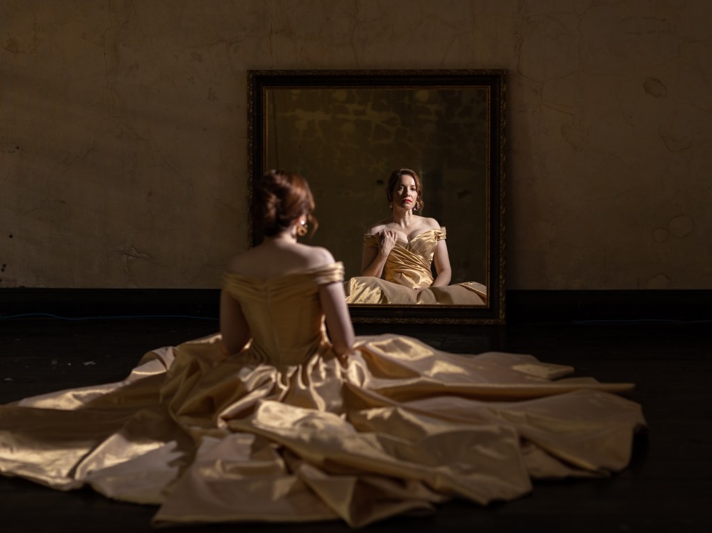 A woman sits with her back to us, an ornate dress spread out on the floor around her; we can see her face reflected in a large mirror sitting opposite her.