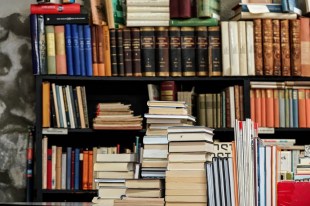 An alluring pile of books and journals sit on a table in front of a crowded bookshelf, tempting the avid reader.