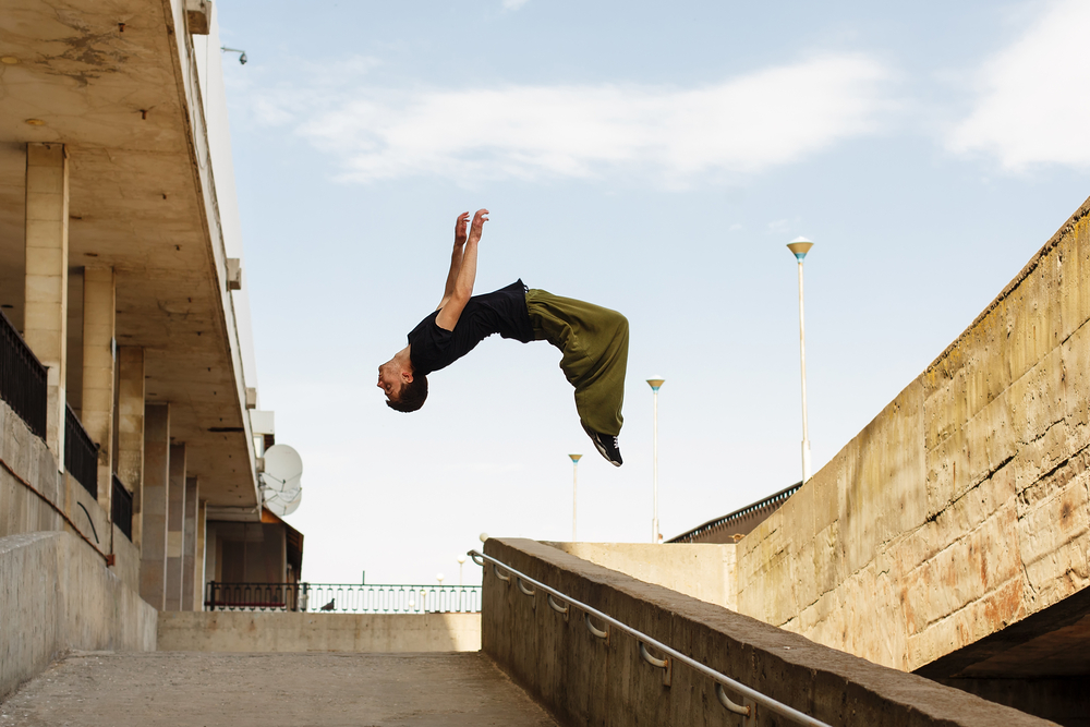 A young man practicing parkour backflips between buildings, silhouetted against the sky.