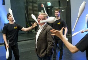 A man wearing glasses smiles as three young jugglers throw their clubs around him.