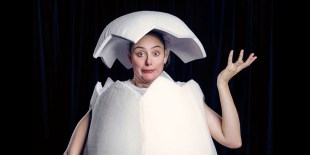 A woman hatches from a a giant egg, with some of the shell worn as a hat. She wears a quizzical expression and holds one hand up to gesture at her egg-hat.