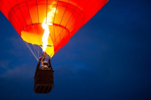 A bright flame generates hot air to lift a brightly coloured hot air balloon against a dark, pre-dawn sky.