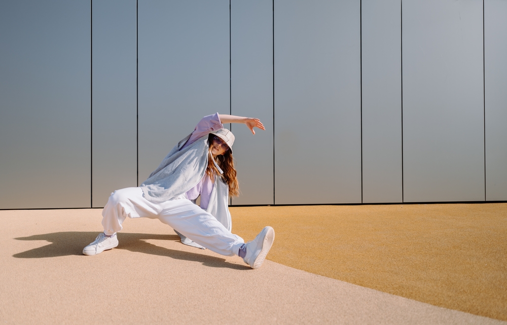 A young b-girl in baggy white clothes and a bucket hat photographed in the midst of a complicated dance move.