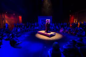 A performer in an elaborate gown and collar stands on a spotlit plinth in a darkened room, surrounded by audience members sitting on the floor.