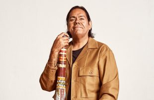 William Barton smiles at the camera. He has shoulder length dark hair and brown eyes. His right hand rests on an ochre-painted didgeridoo and he wears a brown leather jacket over a black t-shirt.