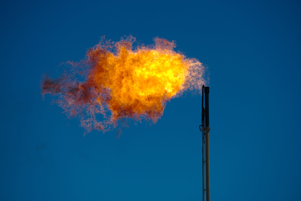 An orange-yellow flare of fire jets from an oil well's stack against a blue sky.