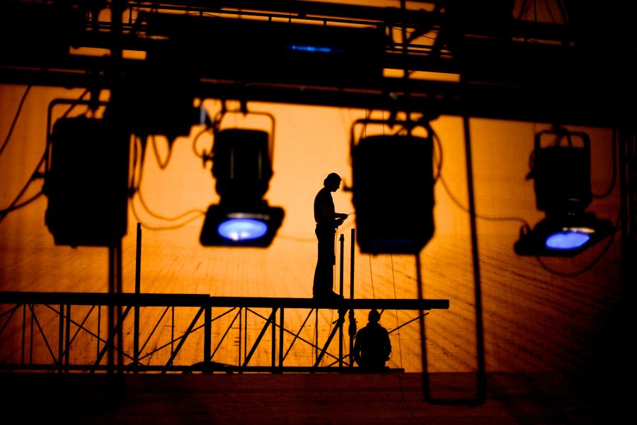 a scene of a darkened area of a theatre backstage, with two production crew members working on the lights, silhouetted by a lit-up background