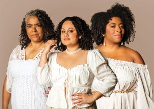 Three dark-skinned women stare at the camera. Each is similarly dressed in white.