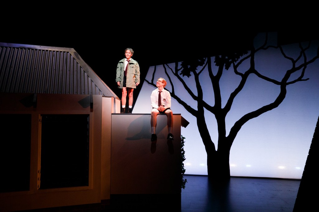 two young performers onstage standing on the roof of a set piece of a house