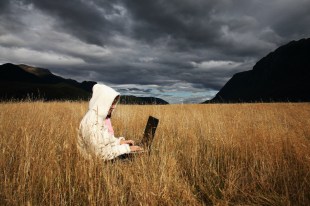 arts news. image is woman wearing white hoodie on laptop in a remote straw field under grey cloudy skies, with black hills in the distance