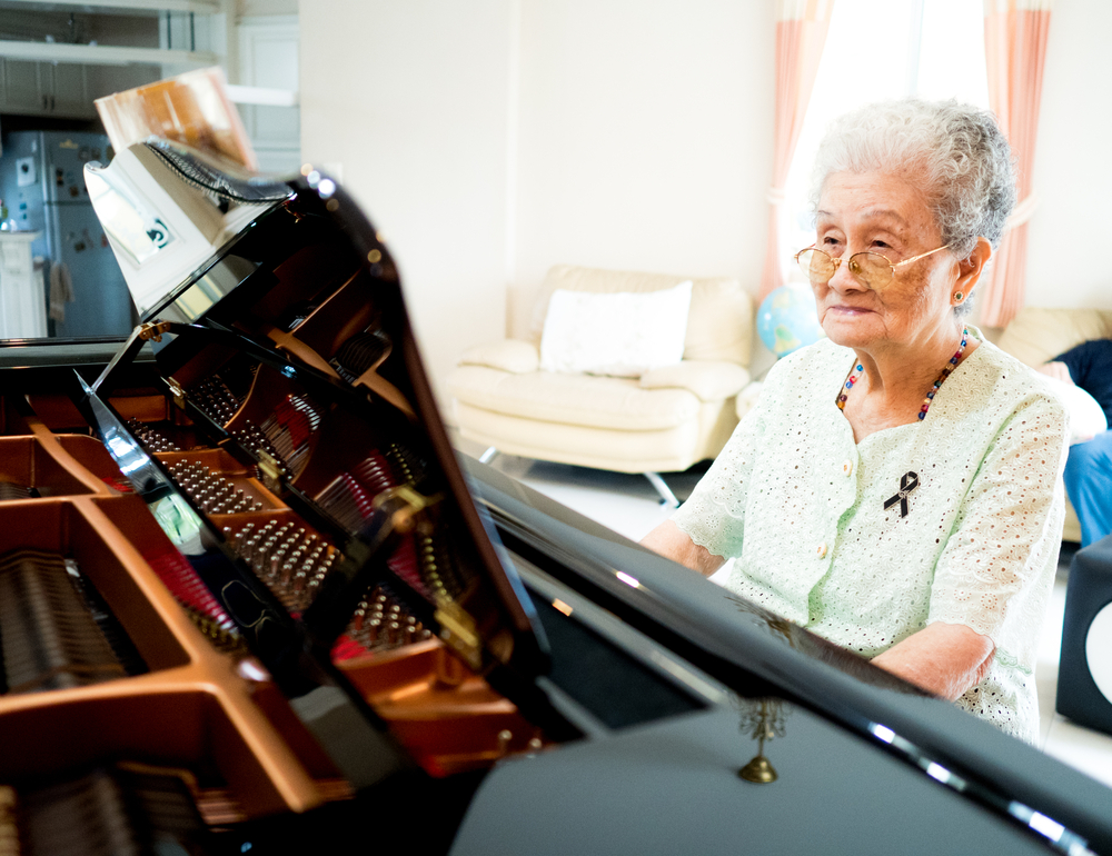 An older woman with grey hair and glasses sits at a piano.