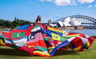 A photograph of Suzy Wrong standing very tall wearing an enormous colourful dress that is made out of national flags stitched together. The dress looks as though it is rippling in the wind. In the background is the Sydney Harbour Bridge and Sydney Opera House.