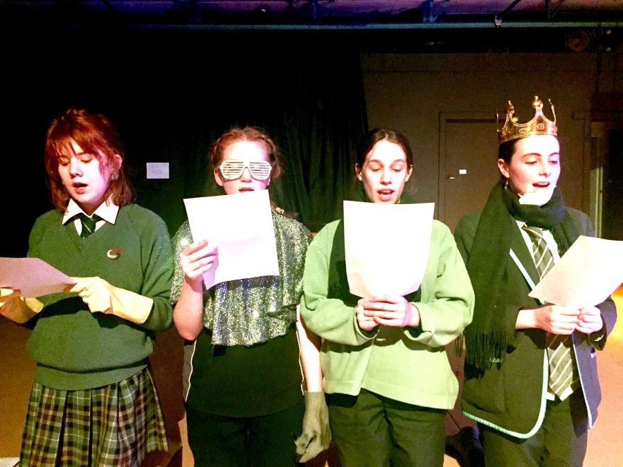 four teenage girls standing in a line reading lines from a play, rehearsing onstage