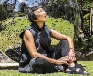 Australia Day Honours. Woman of Asian appearance in sleeveless top sits in front of a bubble like sculpture outdoors looking up toward the sky.