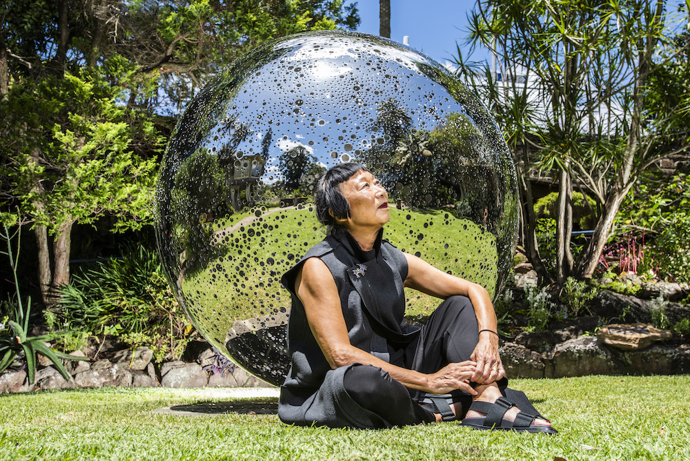 Australia Day Honours. Woman of Asian appearance in sleeveless top sits in front of a bubble like sculpture outdoors looking up toward the sky.
