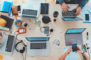 An overhead view of a group of people working on phones and laptops, with a focus on the technology, their hands and assorted coffee cups and notepads.