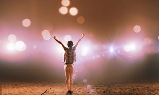 a single female performer facing away from the camera raising their arms in a V, while standing centre stage under a spotlight.